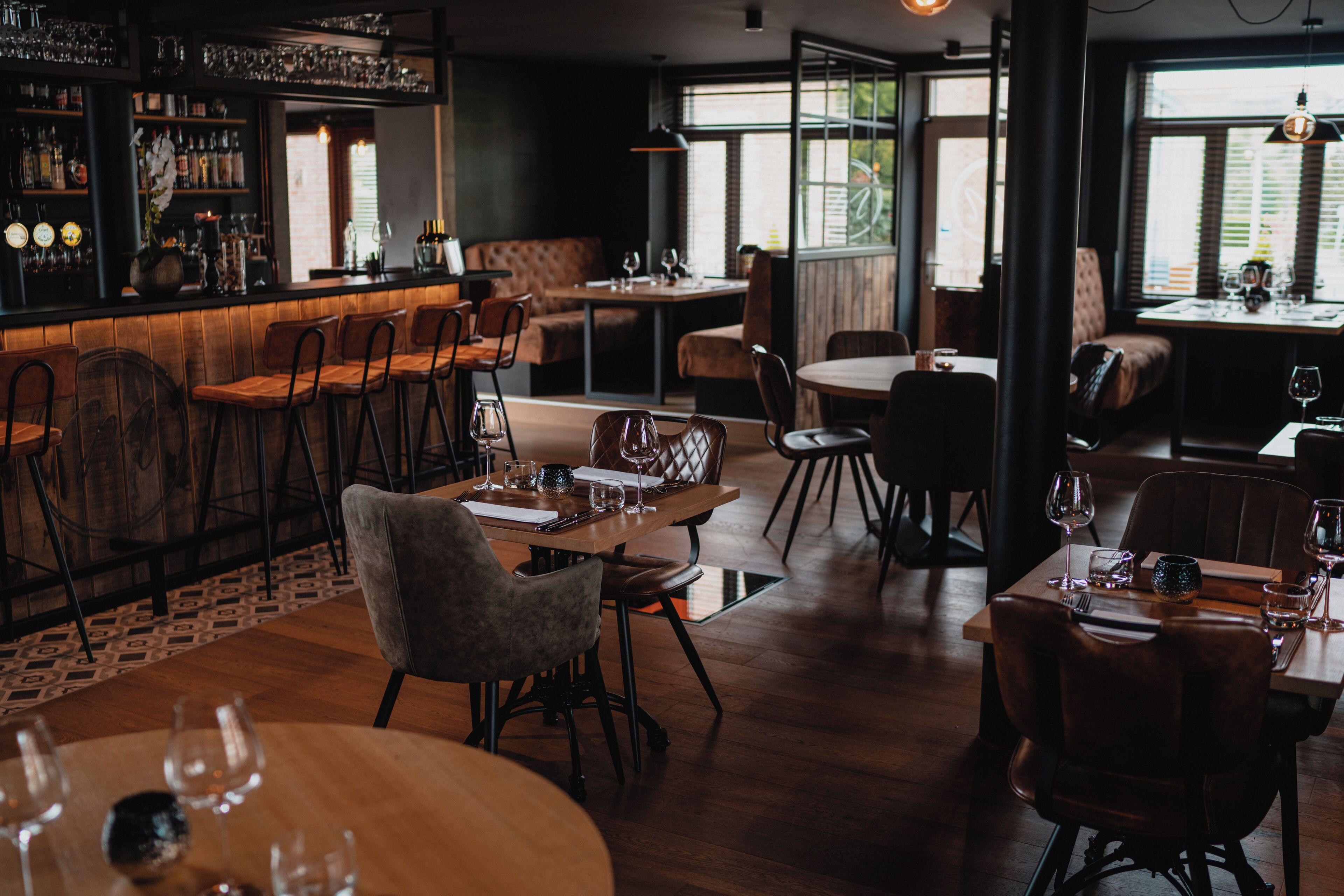 A restaurant interior with wooden tables and chairs, a bar area with shelves of bottles, and large windows allowing natural light.