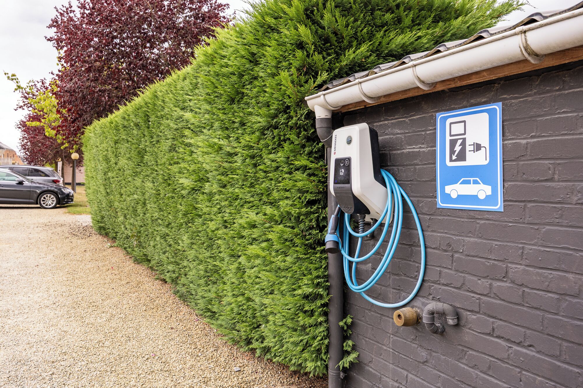 Estación de carga para vehículos eléctricos fijada a la pared, con un letrero azul, un muro de ladrillo y un seto.