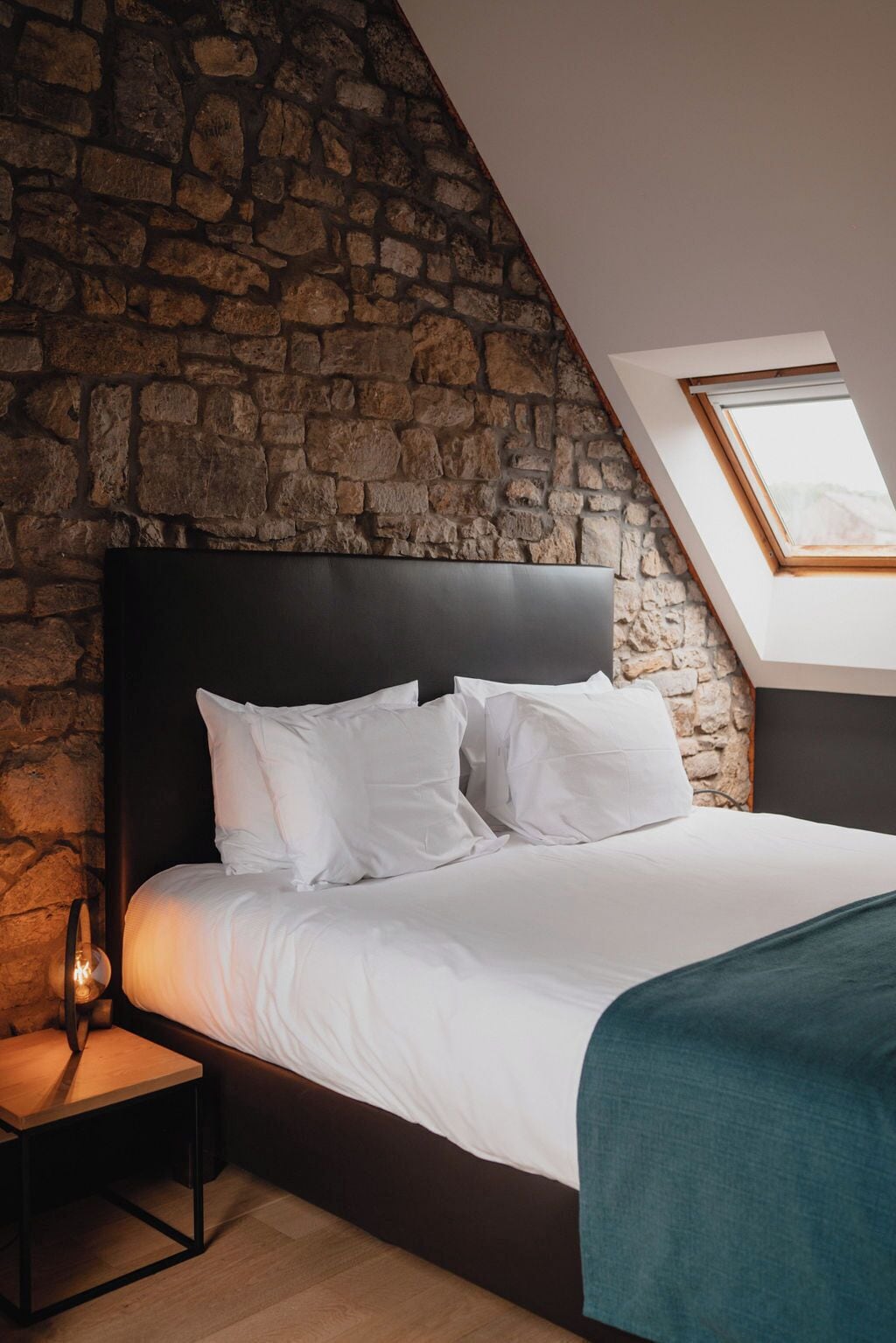 A bedroom with a stone wall, a black headboard, white bedding, a bedside table with a lamp, and a skylight.