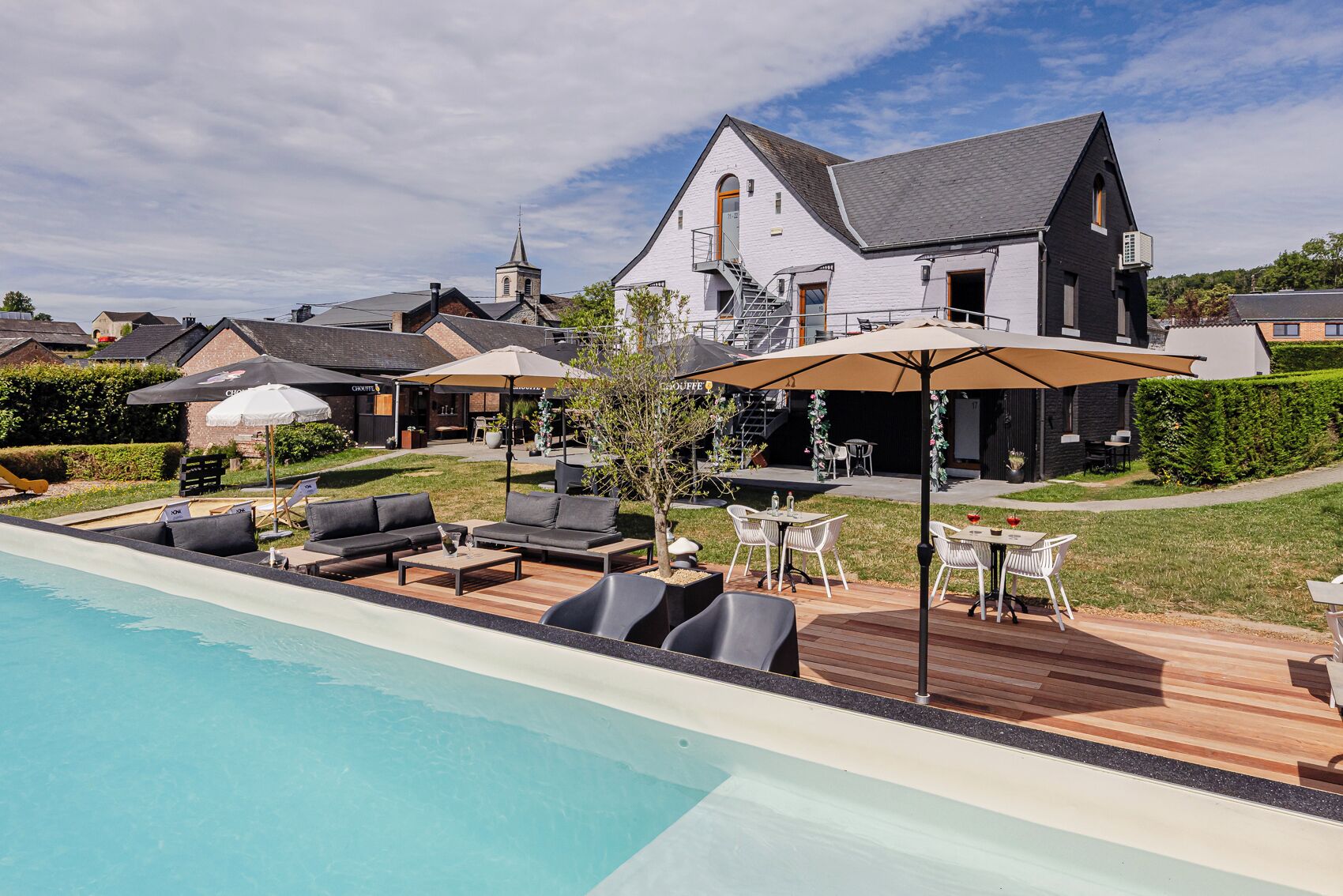 Een zwembad met ligbedden en parasols, een eetgedeelte met tafels en stoelen, en een groot huis met een schoorsteen.