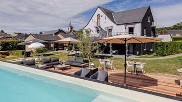 Een zwembad met ligbedden en parasols, een eetgedeelte met tafels en stoelen, en een groot huis met een schoorsteen.