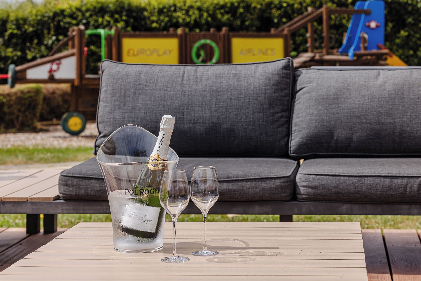 A clear glass bottle of Pol Roger champagne, two empty wine glasses, and a grey sofa in the background.