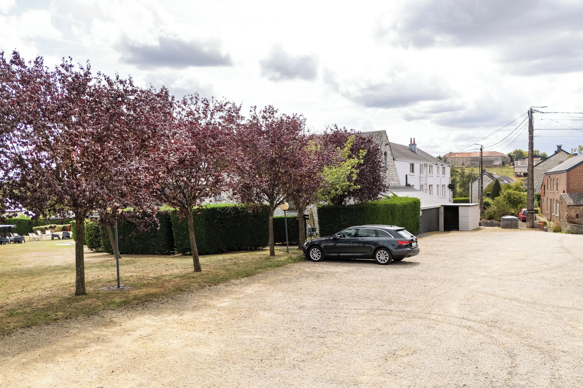 A gravel driveway with a parked car, a tree-lined path, and residential buildings in the background.