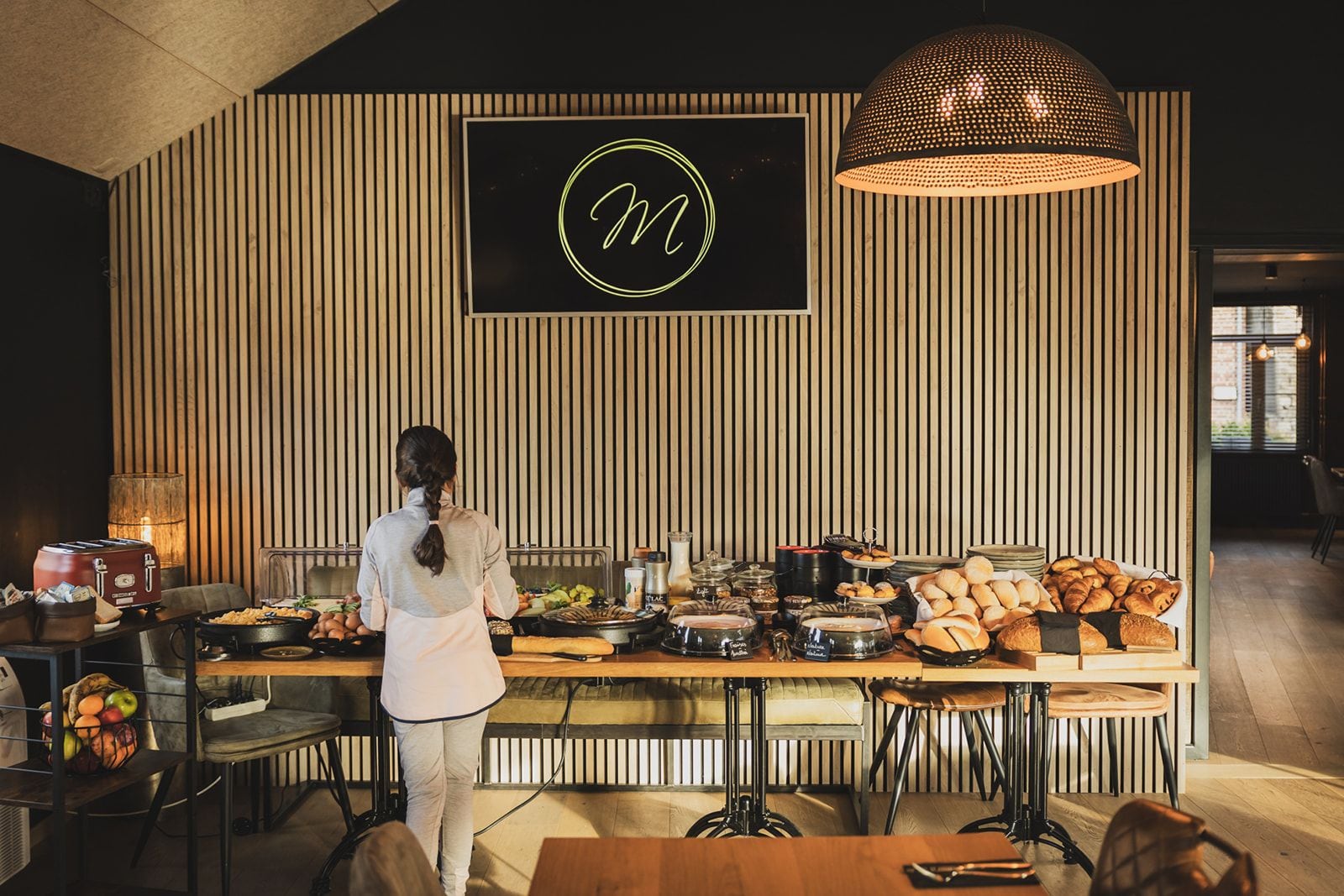 Montaje de buffet con diversos alimentos, una persona sirviendo y un letrero iluminado grande en la pared.