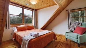 A bedroom with a wooden ceiling, a bed with orange bedding, a green armchair, and a view of trees through the windows. - Secluded & Tranquil Retreat for 14 in the Poconos – Firepit/Games/Stargazing+ (Lake Ariel)
