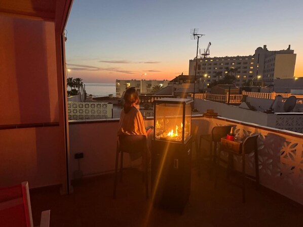 Un balcon doté d’un espace fermé par des parois en verre, comprenant un coin salon et offrant une vue sur la mer et les bâtiments au coucher du soleil.