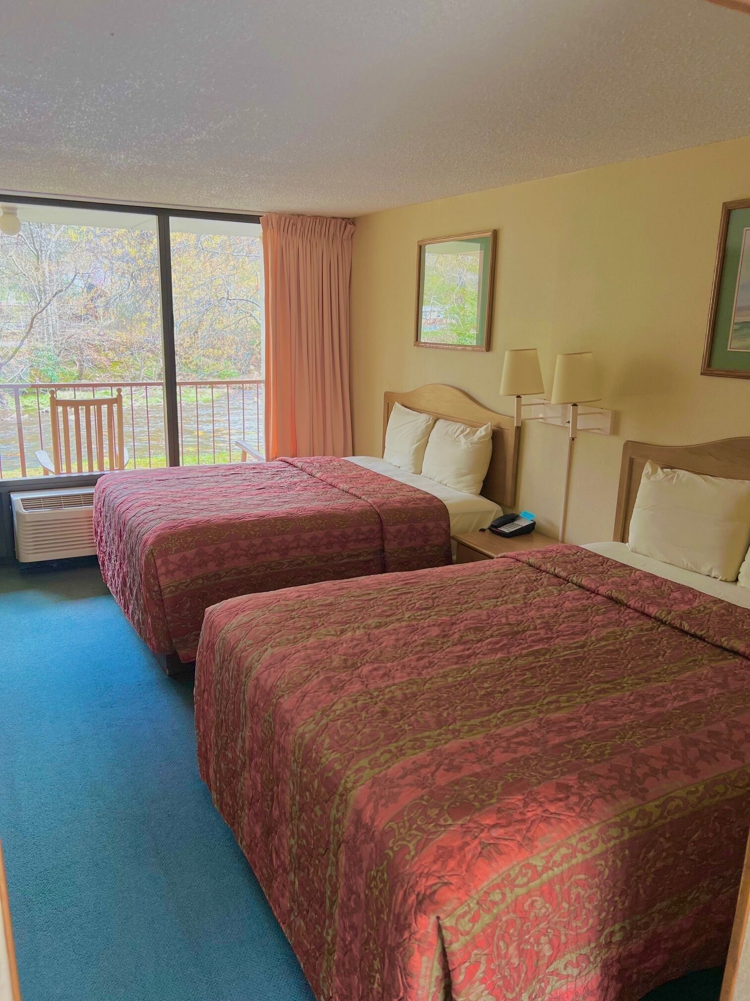 A hotel room with two beds, a window with a view of trees, a chair, and a nightstand with a lamp.