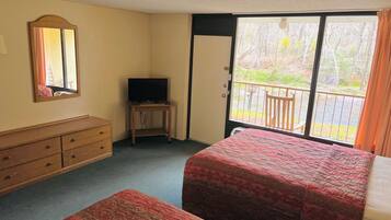 A hotel room with two beds, a dresser, a television on a small table, and a balcony with a view of trees.