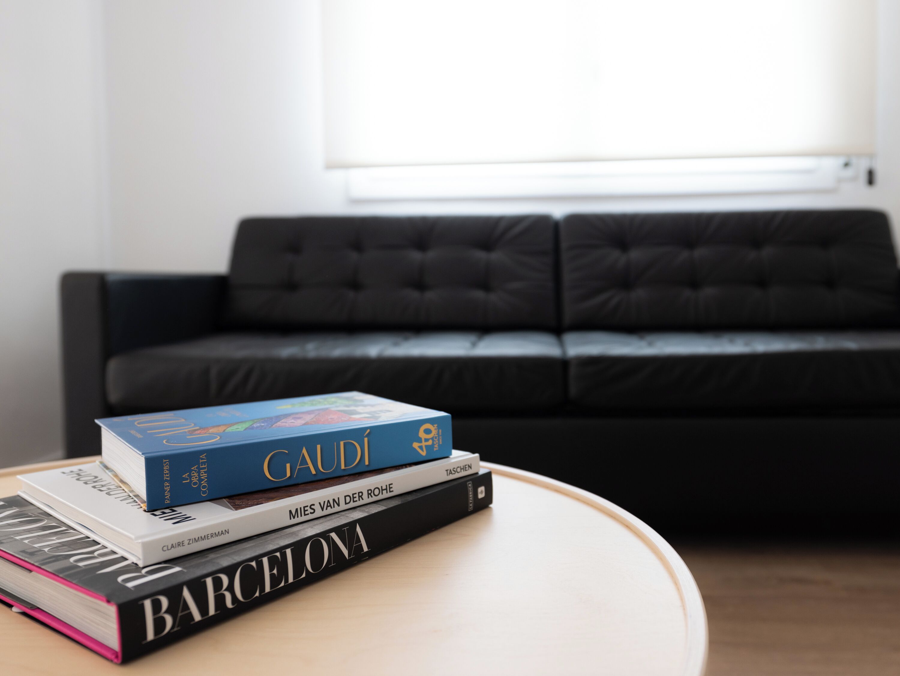 A stack of books on a round table with a black leather sofa in the background.
