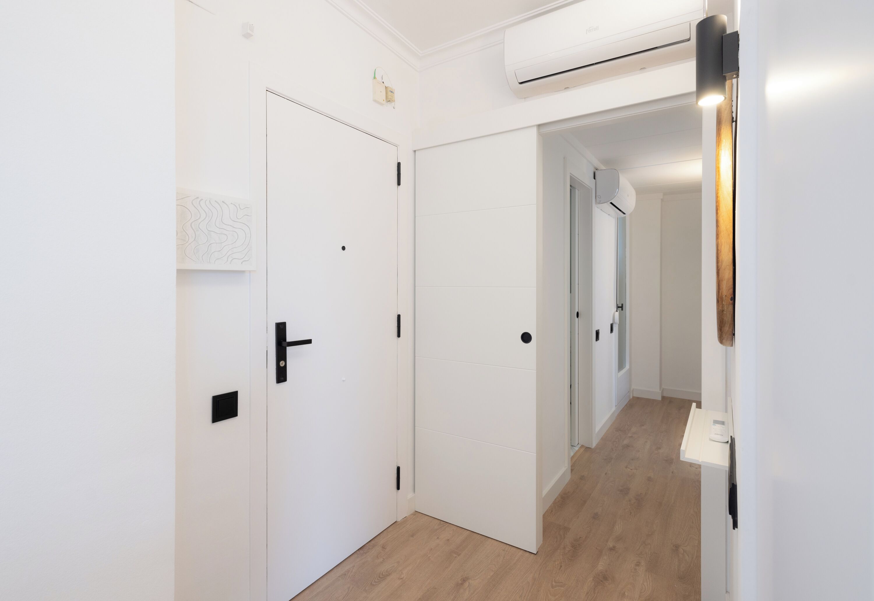 Un pasillo con paredes blancas y piso de madera, con una puerta blanca, un picaporte negro y un pequeño cuadrado negro en la pared.
