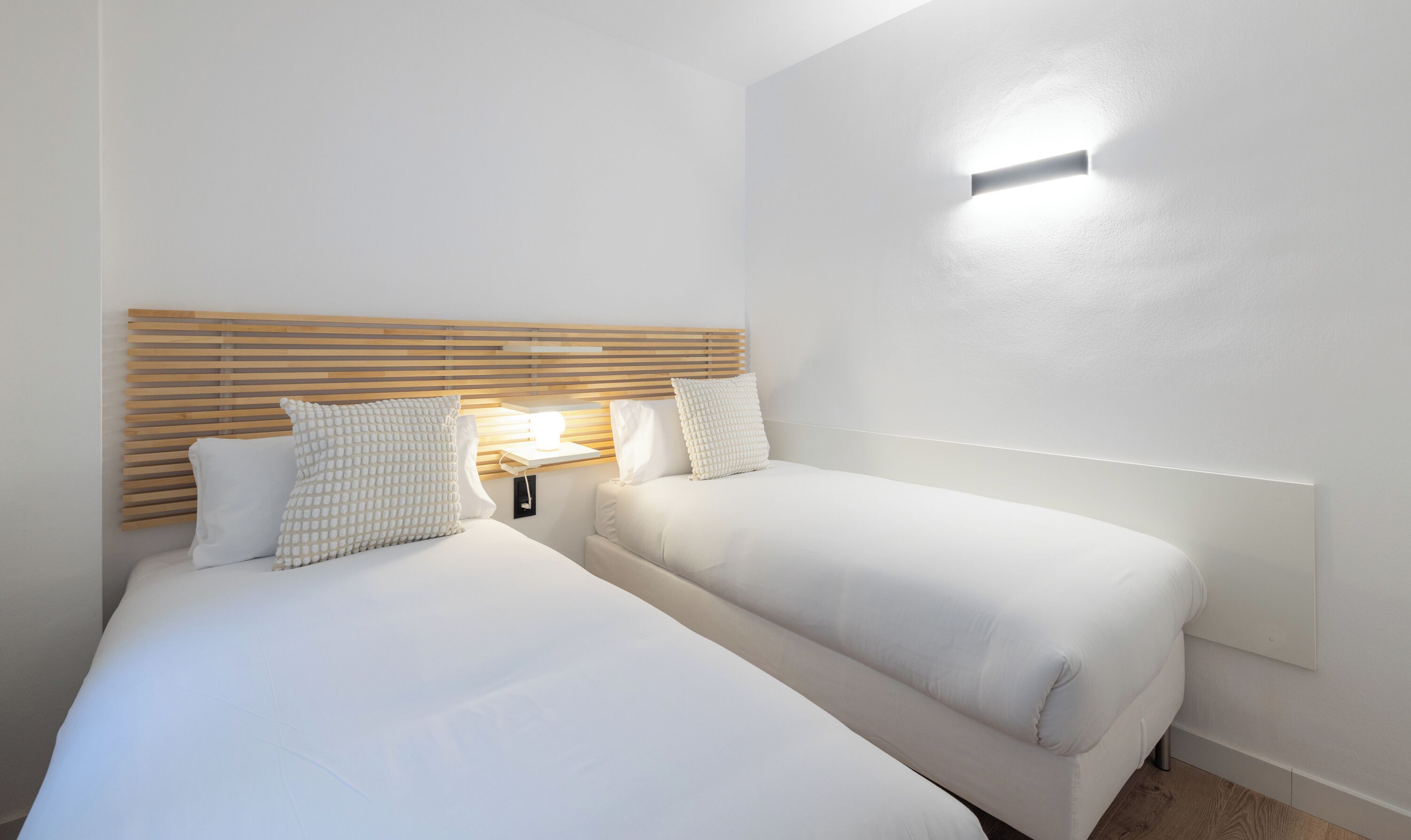 Habitación de hotel con dos camas, cabecero de madera, mesita de noche con lámpara y una lámpara de pared.