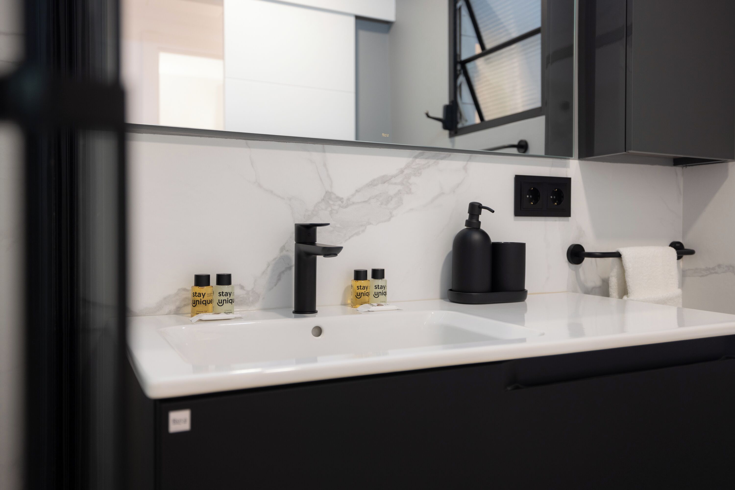 Une salle de bain moderne avec un comptoir en marbre blanc, des accessoires noirs et deux flacons de savon liquide aux parfums originaux.