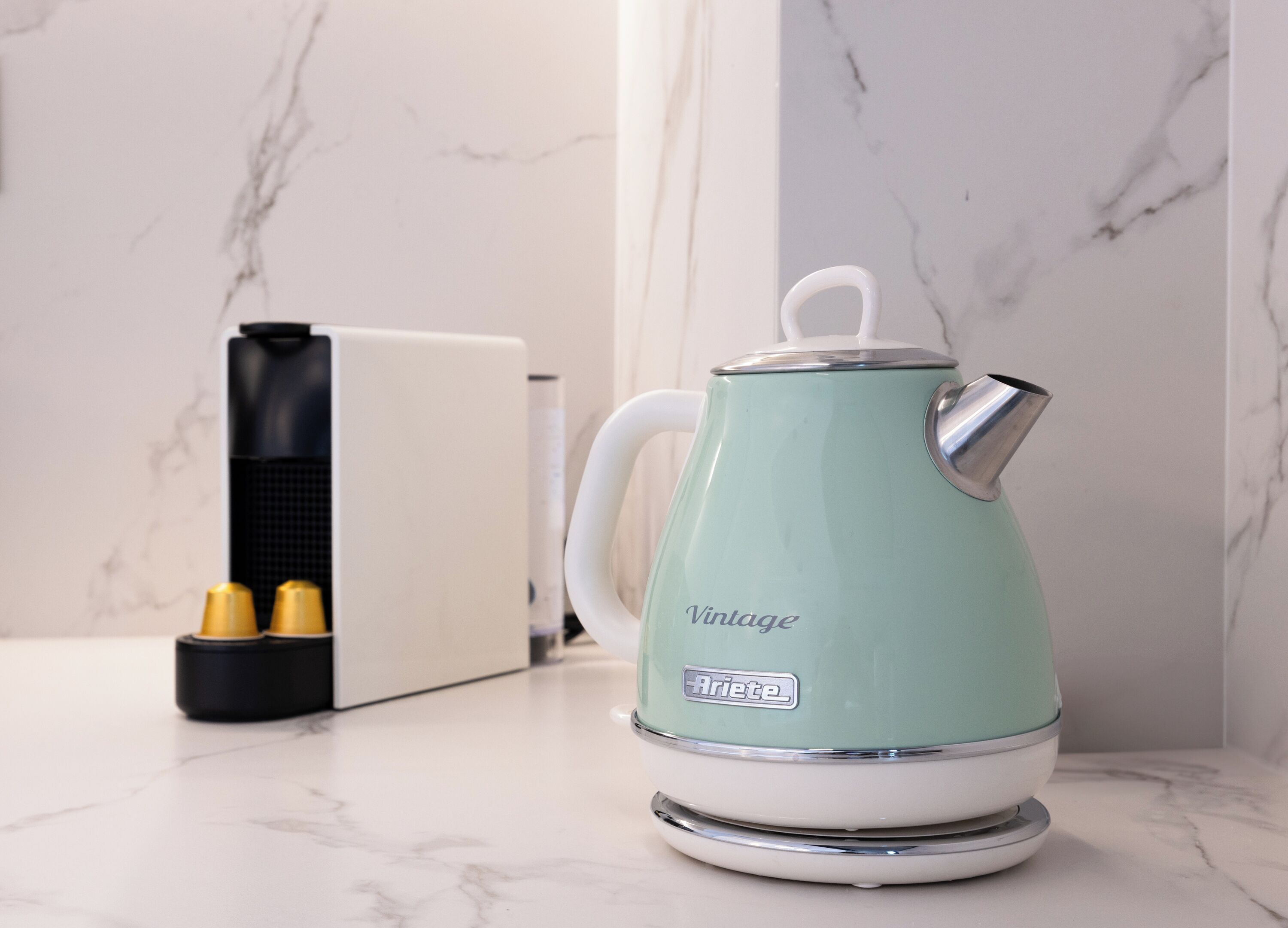 A vintage-style kettle on a marble countertop with a modern coffee maker and two yellow cups in the background.