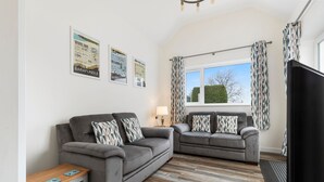 A living room with two grey sofas, a wooden coffee table, and a TV. There are patterned curtains and a window with a view of trees. - Fern Cottage - Dog Friendly Cottage with Hot Tub (Begelly)