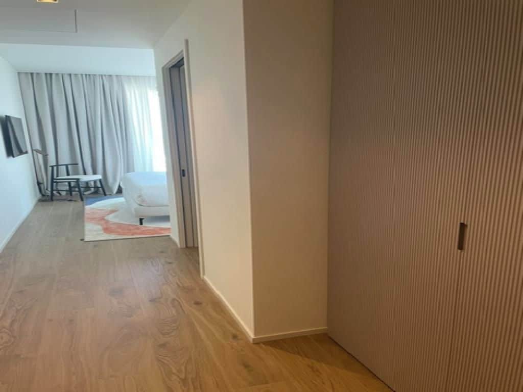 A hallway with a wooden floor, a white door, and a room in the background with a white chair and a small table.