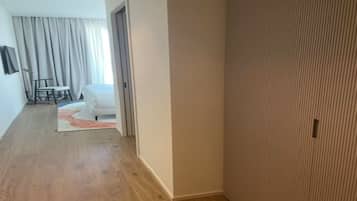 A hallway with a wooden floor, a white door, and a room in the background with a white chair and a small table.