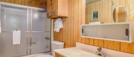 Un baño con paneles de madera, mampara de ducha de vidrio, inodoro, lavabo con espejo y toallero.