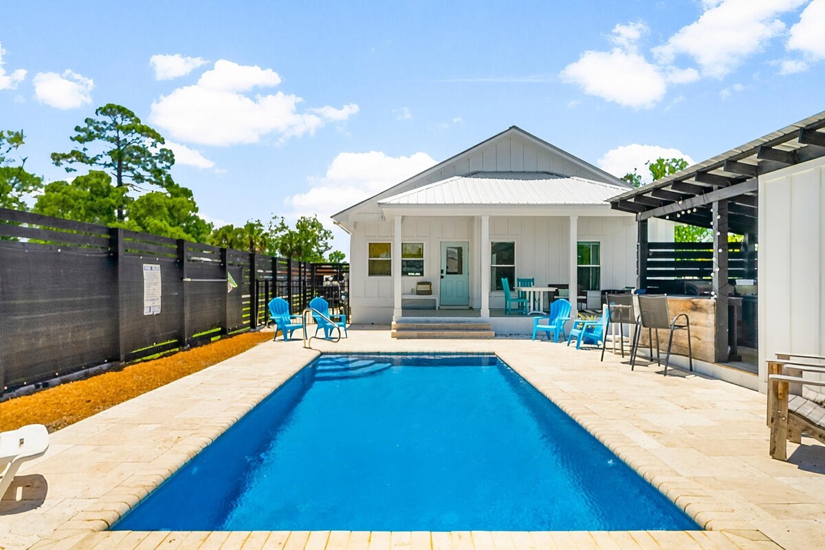 A rectangular swimming pool with a white house in the background, surrounded by a fence and outdoor seating.