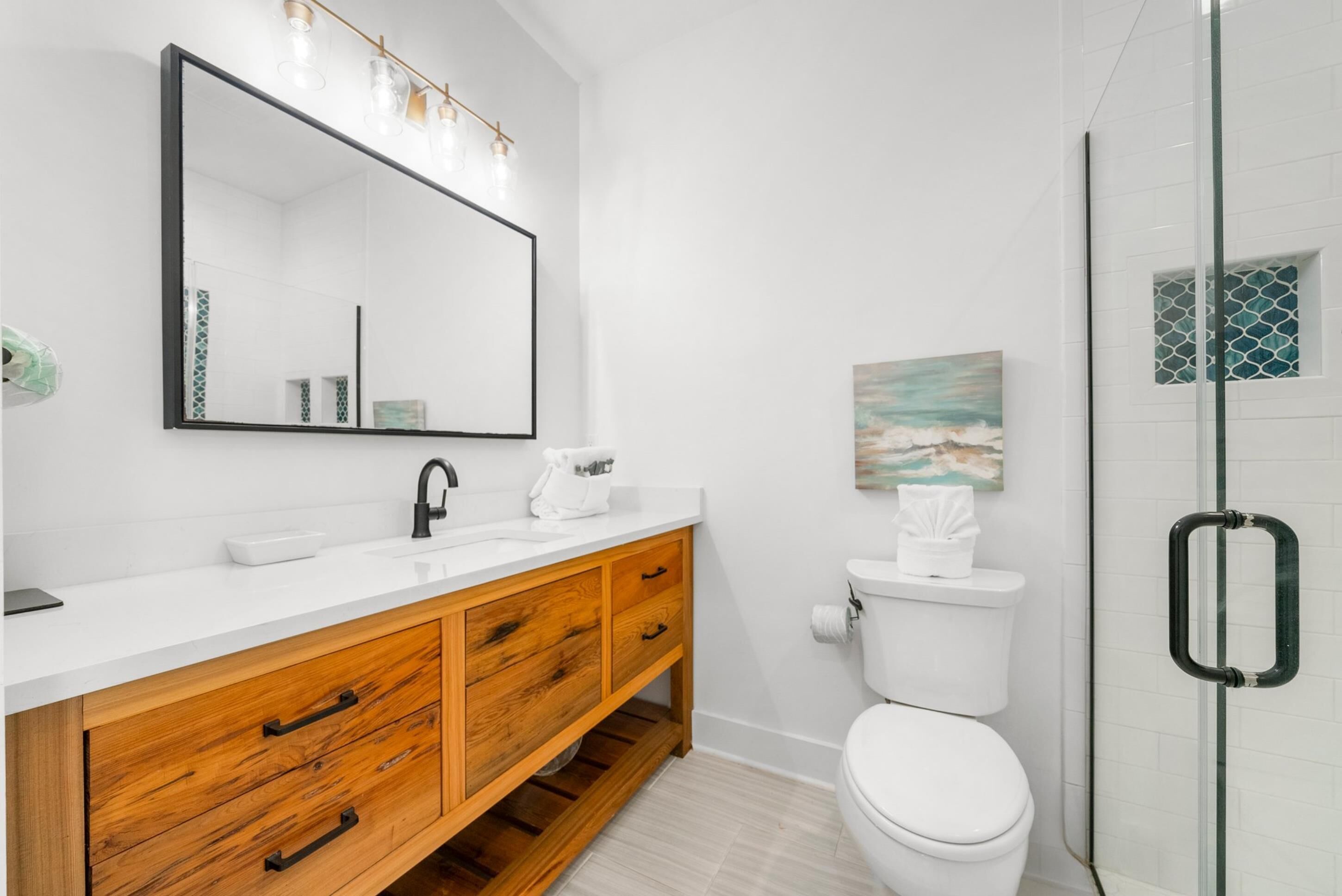 A modern bathroom with a white sink, wooden vanity, a large mirror, a glass shower enclosure, and a painting on the wall.