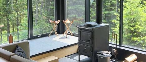 A modern interior with a large window, a sofa, a fireplace, and a view of the forest.