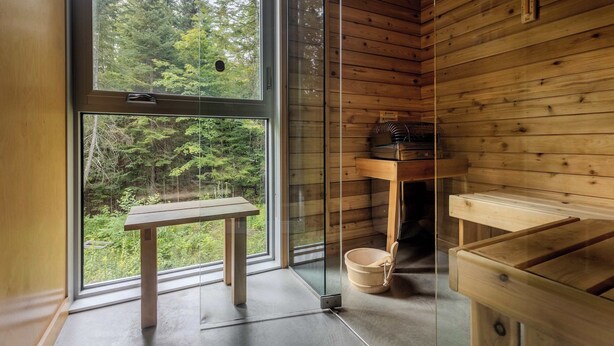 Een houten hutje interieur met een groot raam, een houten bank, een glazen douchecabine en een houten plank met spullen.