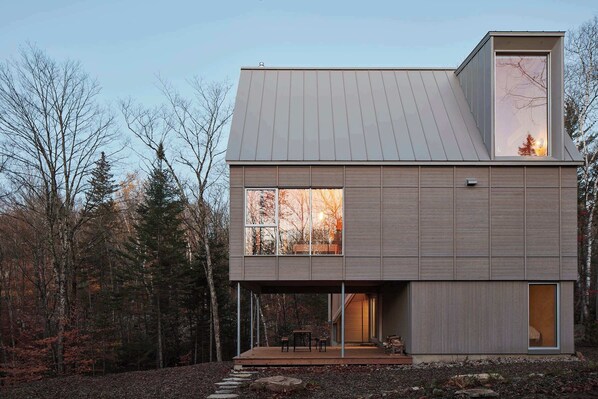 A modern two-story house with a large window, a deck, and a forested backdrop. - The Shoal (Lanaudière)