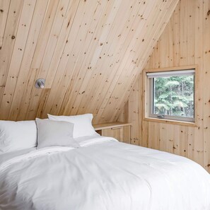 A bedroom with a sloped ceiling, a window with a view of trees, and a bed with white bedding.