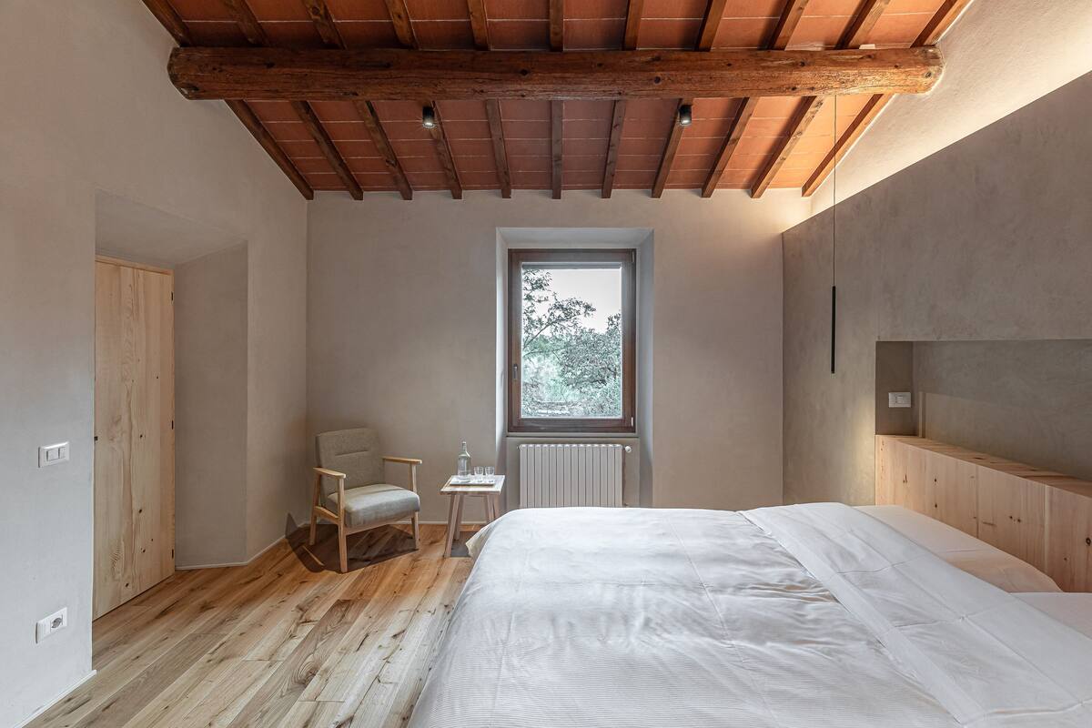 Camera da letto con soffitto in legno, una finestra vista alberi, un letto, una sedia e un tavolino con un bicchiere.