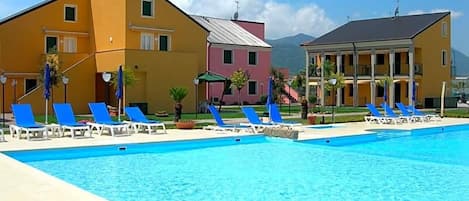 A swimming pool with blue lounge chairs, a yellow building, and a pink building in the background.