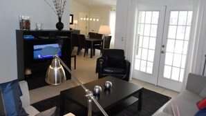 A living room with a TV, black coffee table, and a lamp. There is a dining area in the background with chairs and a table.