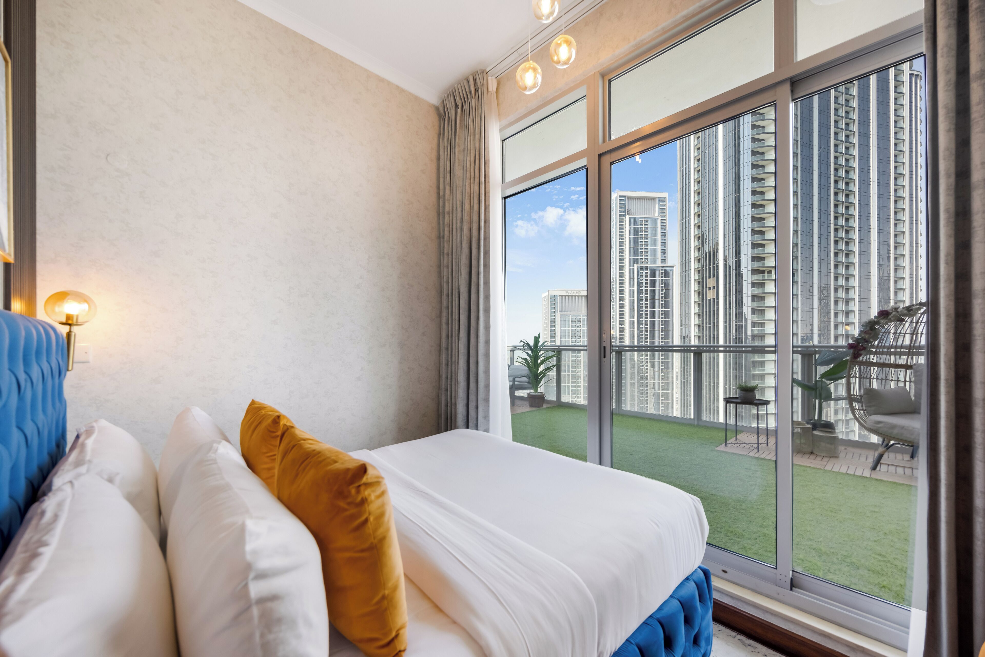 A bedroom with a large window, a bed with white and blue bedding, and a view of high-rise buildings.