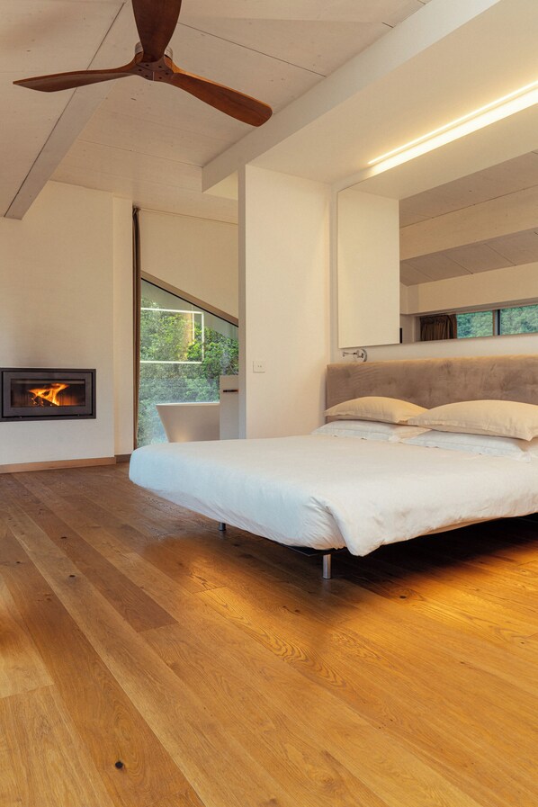 A modern bedroom with a large bed, wooden flooring, a ceiling fan, a fireplace, and a view of greenery through a window. - LA FORESTALE Luxury Ecolodge (Acqualagna)