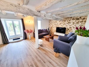 A modern living room with a stone wall, a flat-screen TV, a grey sofa, a coffee table, and a large window with curtains. - Nature View Cottage • Terrace • Piscina (Aguts)