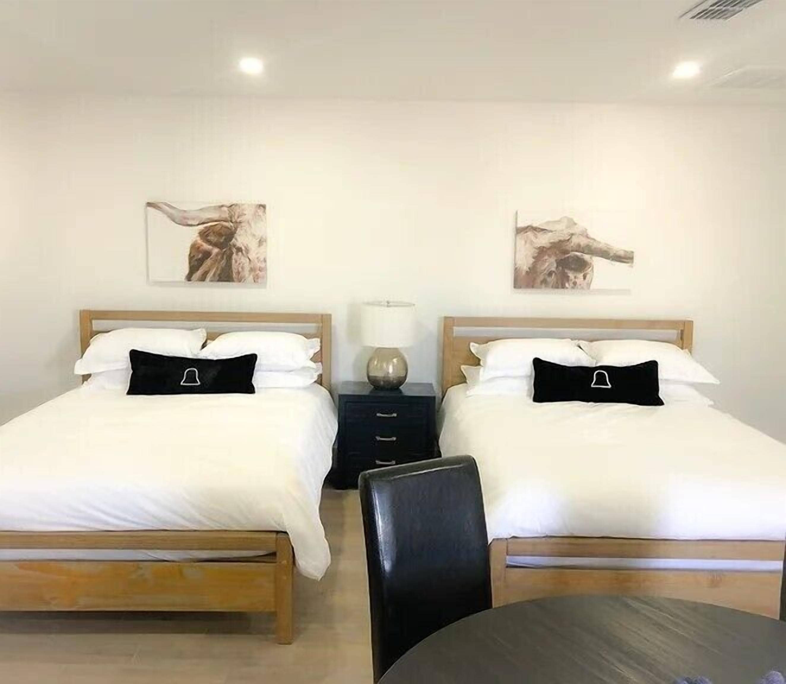A hotel room with two beds, each with white bedding and black pillows, a wooden headboard, a small bedside table with a lamp, and two wall-mounted paintings.