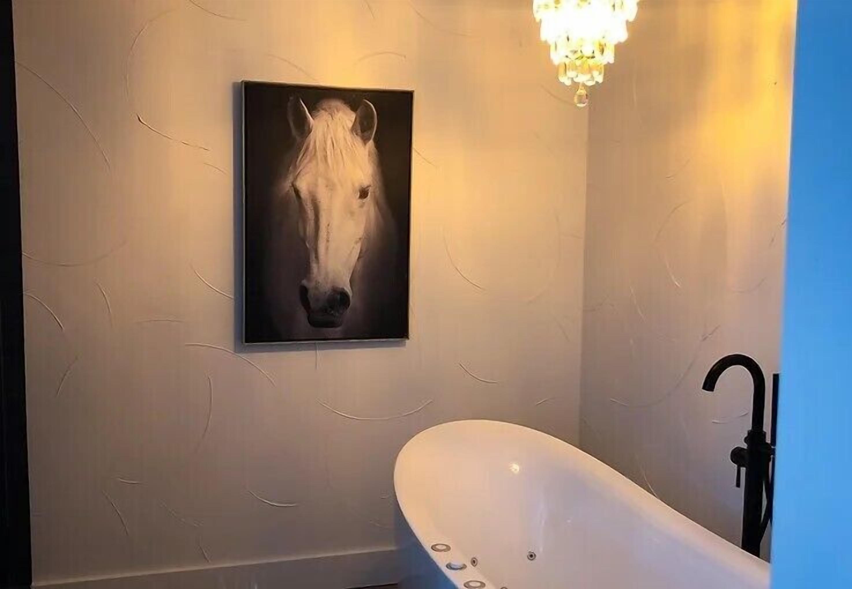 A bathroom with a white bathtub, a wall-mounted horse painting, and a chandelier.