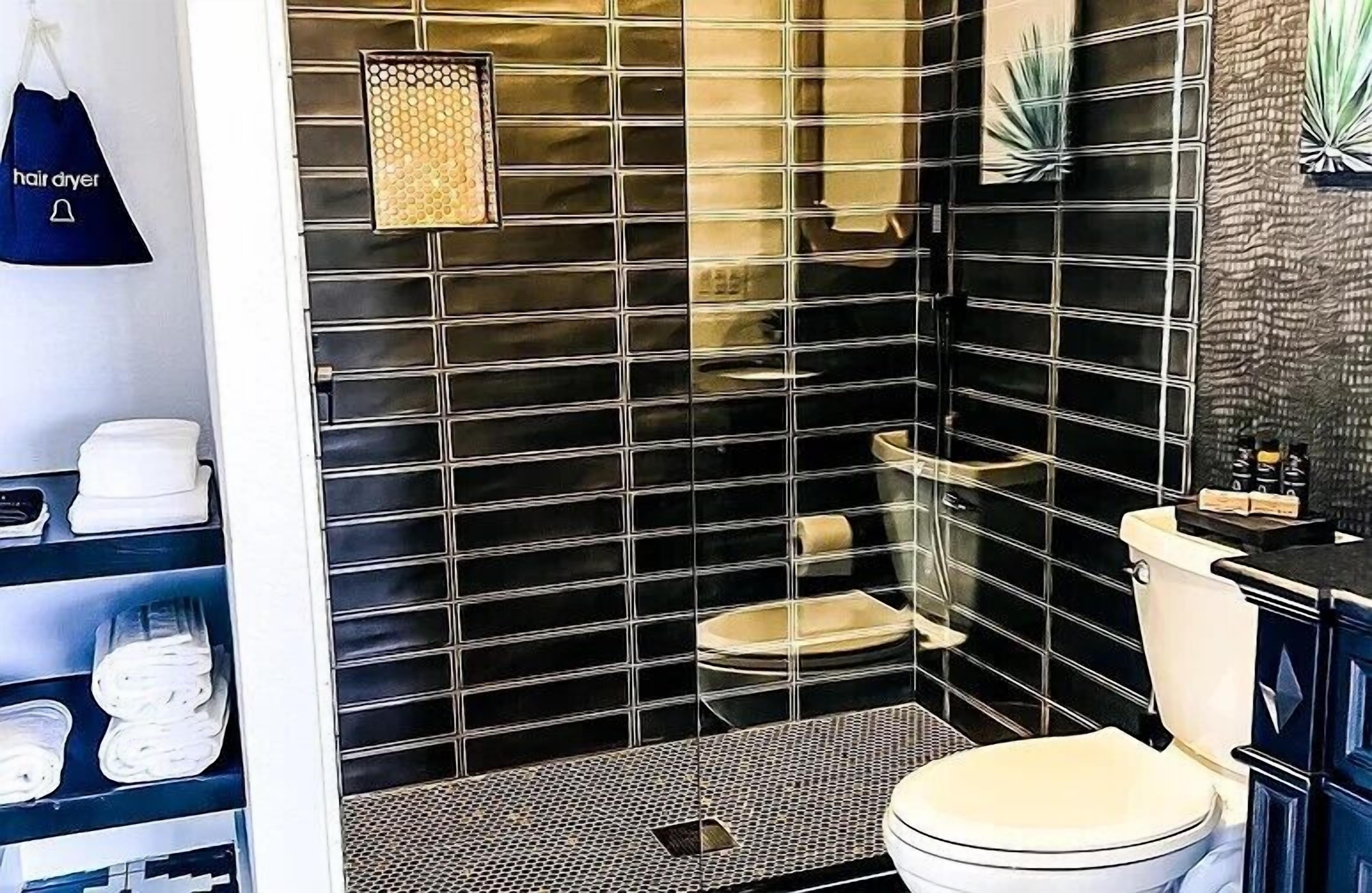 Un baño moderno con paredes de azulejos negros, un inodoro blanco, una zona de ducha y un estante con secadores de pelo.