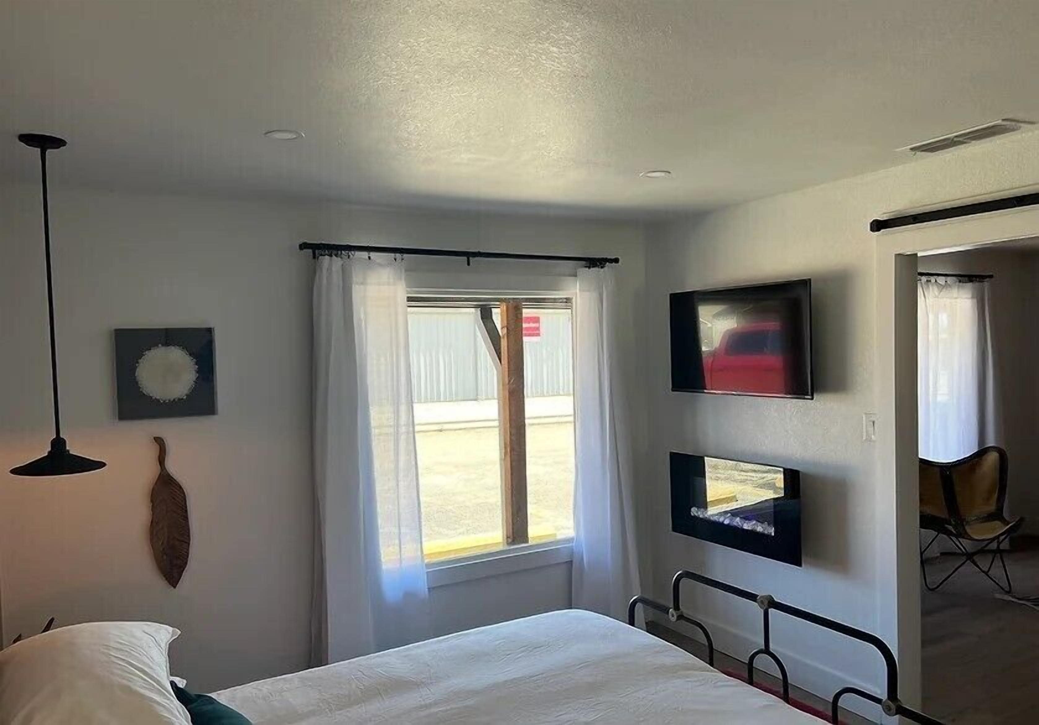 Habitación de hotel con cama, televisor de pantalla plana en la pared, ventana con cortinas, una silla y una obra de arte colgada en la pared.