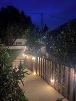 Un sendero bordeado por una valla de madera con luces uniformemente distribuidas, que conduce a un edificio con un banco visible y un árbol al fondo.