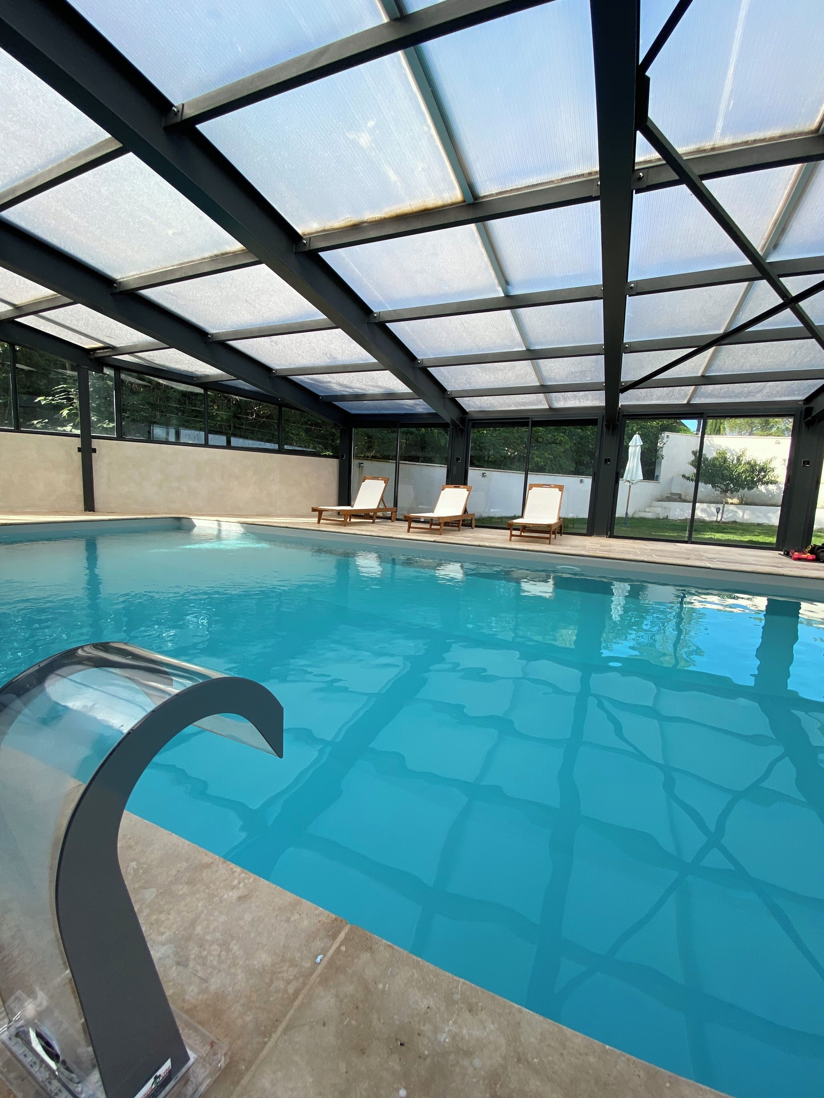 A swimming pool with a glass ceiling, clear blue water, and three lounge chairs.