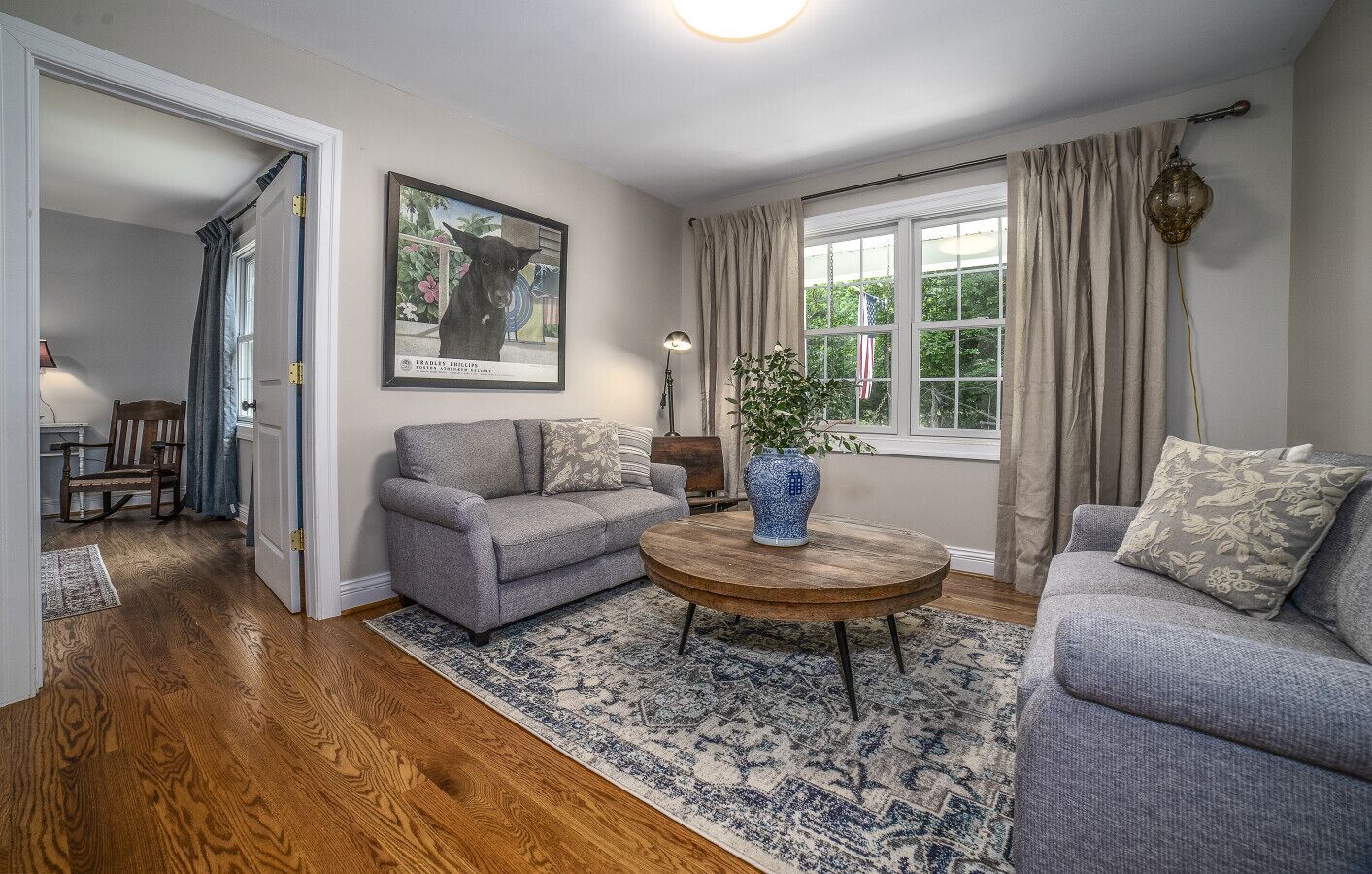 A living room with a grey sofa, a round wooden coffee table, and a blue vase. There is a painting on the wall and a window with curtains.