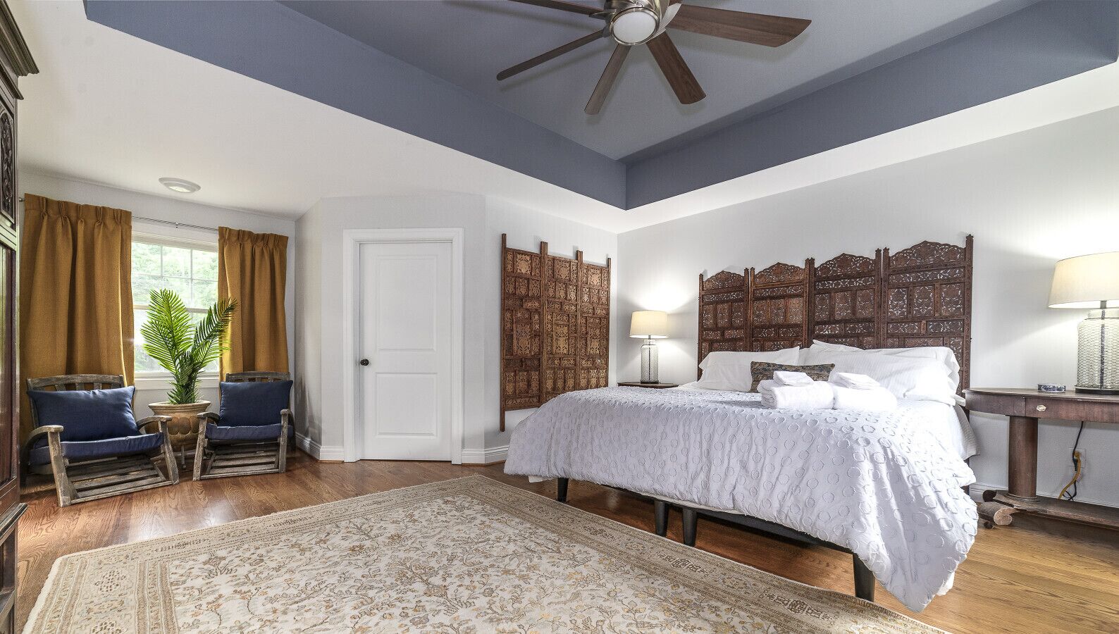 A bedroom with a large bed, a ceiling fan, a wooden headboard, a nightstand, a lamp, and a potted plant.