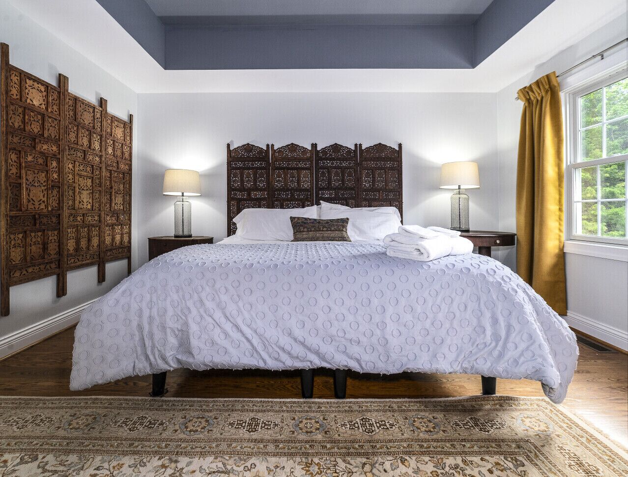 A bedroom with a large bed, wooden headboard, two bedside tables with lamps, a patterned rug, and a view of greenery through a window with yellow curtains.