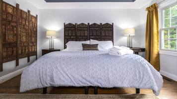 A bedroom with a large bed, wooden headboard, two bedside tables with lamps, a patterned rug, and a view of greenery through a window with yellow curtains.