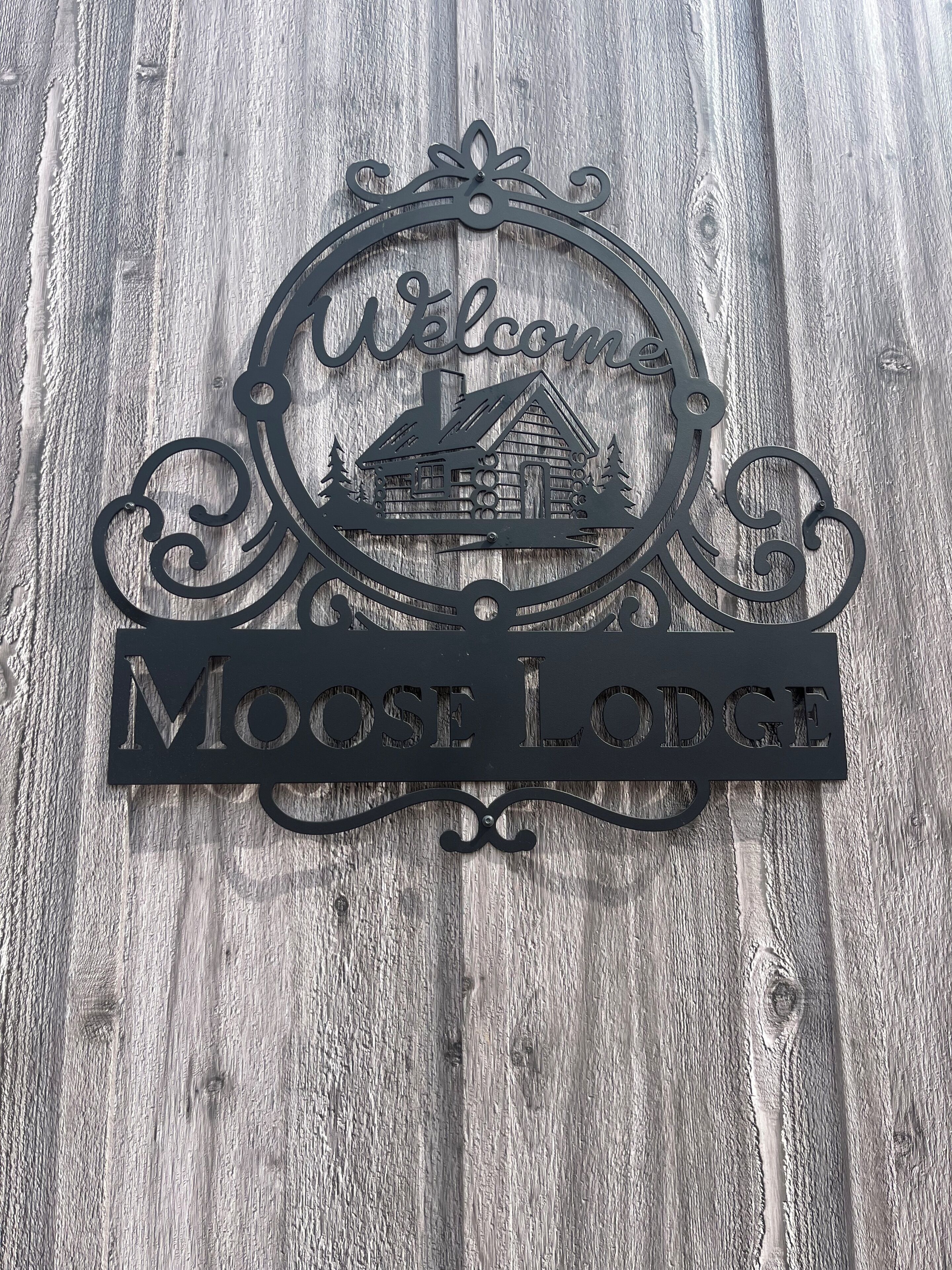 Un letrero de metal negro con diseño rústico, que muestra una casa y árboles, y la inscripción 'Welcome Moose Lodge'.