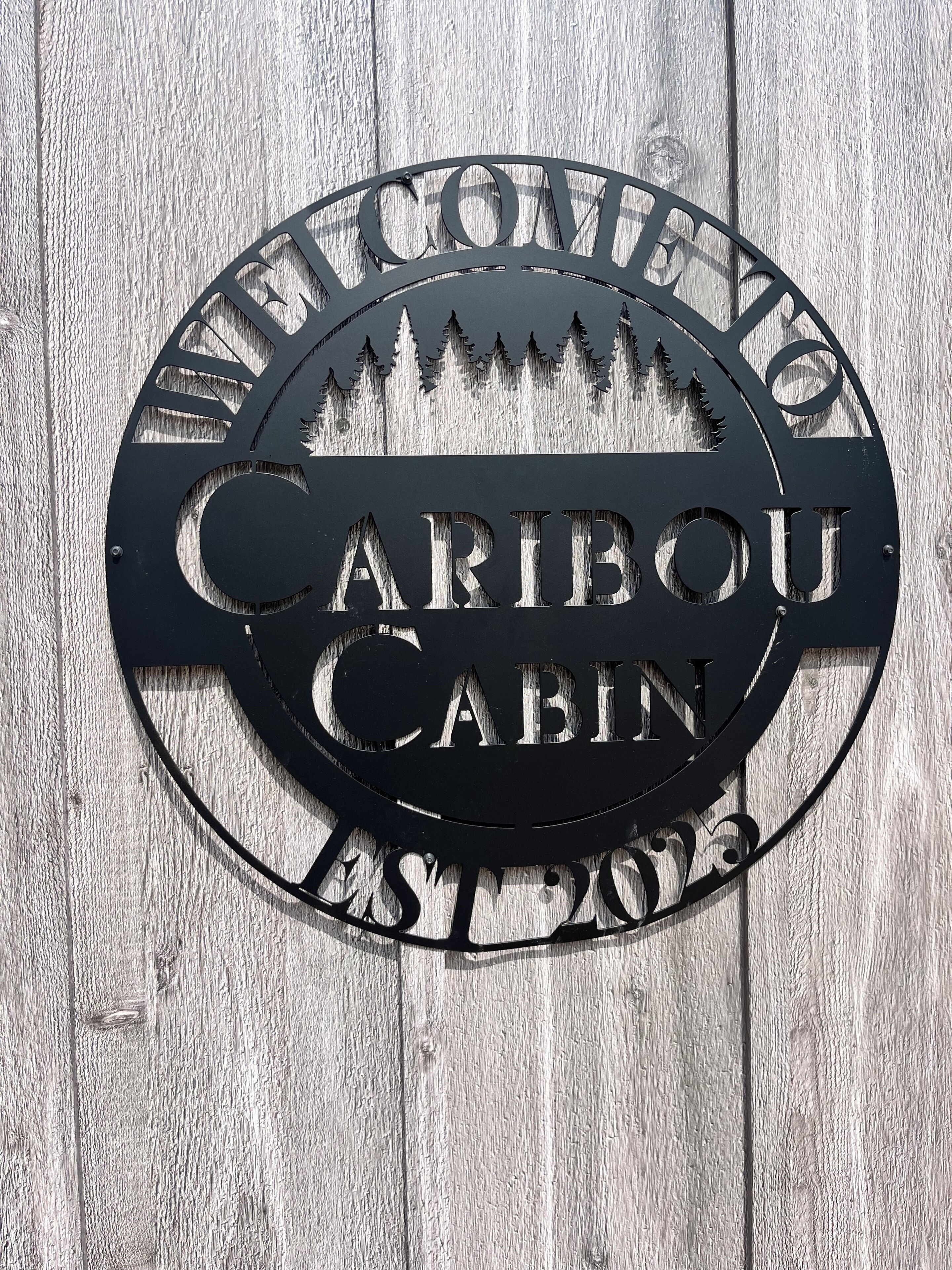 A metal sign with the text "WELCOME TO CARIBOU CABIN EST. 2013" against a wooden background.