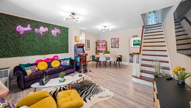 Ein modernes Wohnzimmer mit einer grünen Wand, einem violetten Sofa und einem Neon-Leuchtschild.