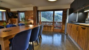 Eine moderne Küche mit Holzschränken, ein Essbereich mit blauen Stühlen und ein großes Fenster mit Blick auf eine gebirgige Landschaft.
