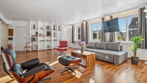 Ett modernt vardagsrum med en grå soffa, en röd fåtölj och ett soffbord i trä. Det finns en bokhylla med diverse föremål och en platt-TV som är monterad på väggen.