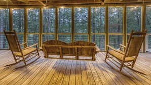 A wooden deck with a swing chair and two rocking chairs, large windows with a forest view. - Pet-Friendly Creekside Cabin w/ Hot Tub 2BR/2 Bath (Ellijay)
