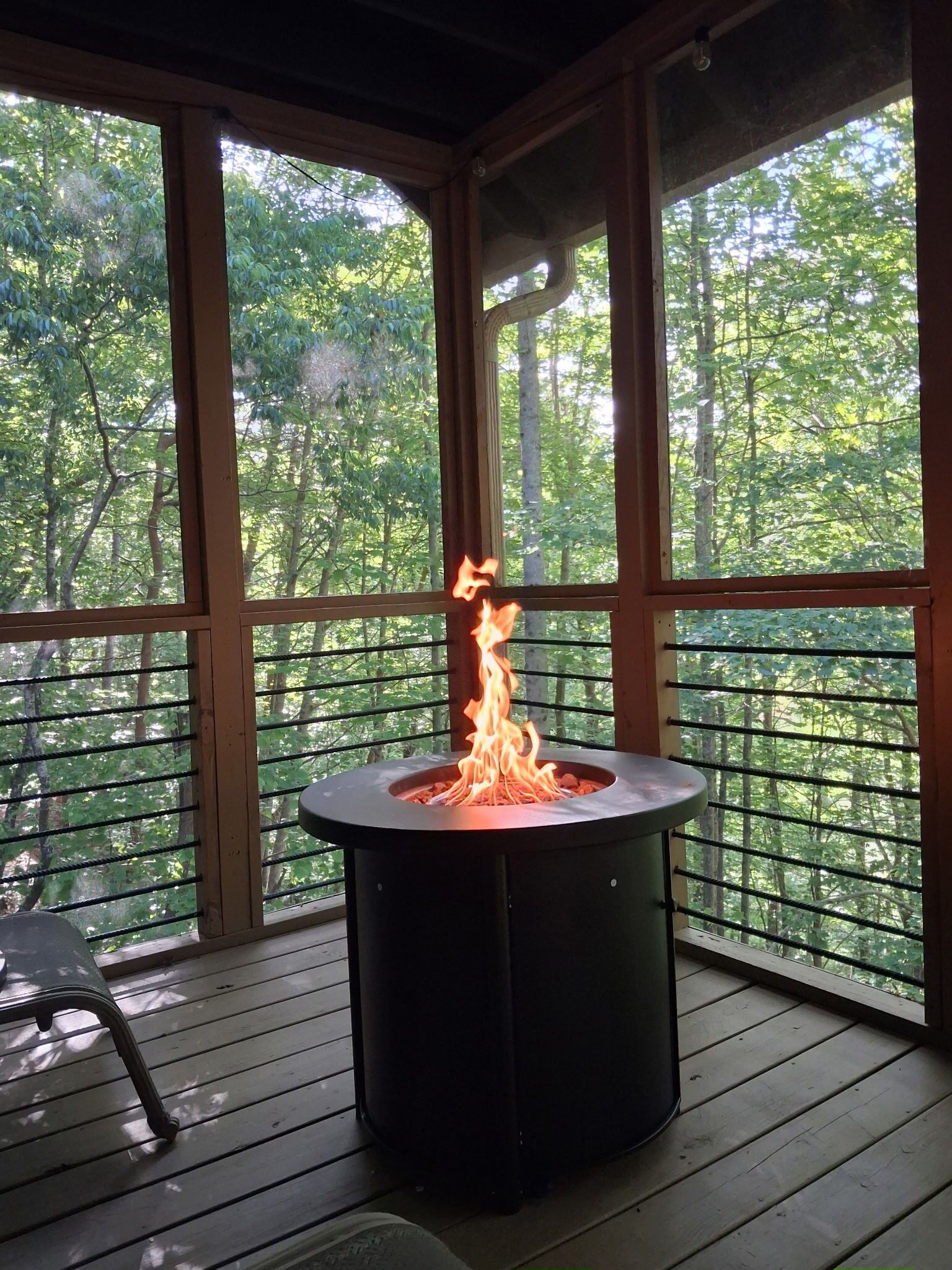 Un pozo de fuego circular al aire libre con llamas, rodeado por una terraza de madera y amplias ventanas con vistas a un área boscosa.